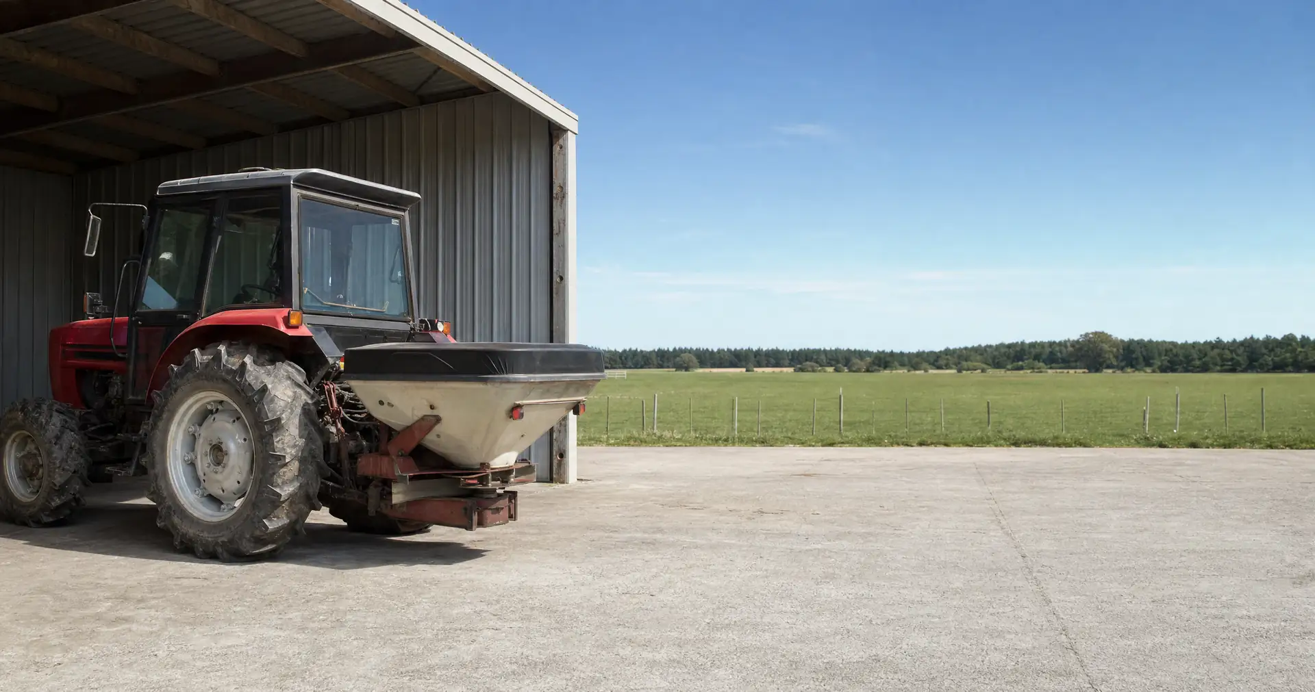 A tractor with a mounted fertiliser spreader parked on the apron of an open-bay implement shed