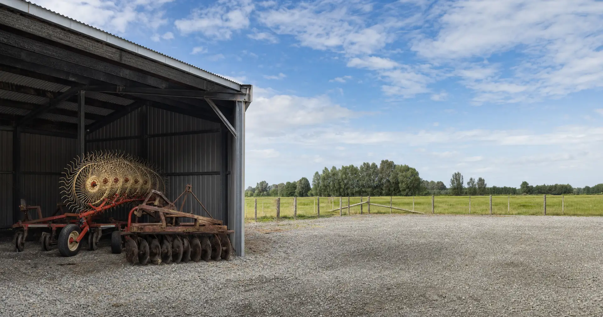 An open-bay implement shed with a hay rake and a set of discs parked under cover